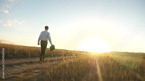 businessman in sunglasses goes down country road with briefcase in his hand. agricultural business concept. The entrepreneur works in a rural area. farmer inspects the land at sunset. Slow motion