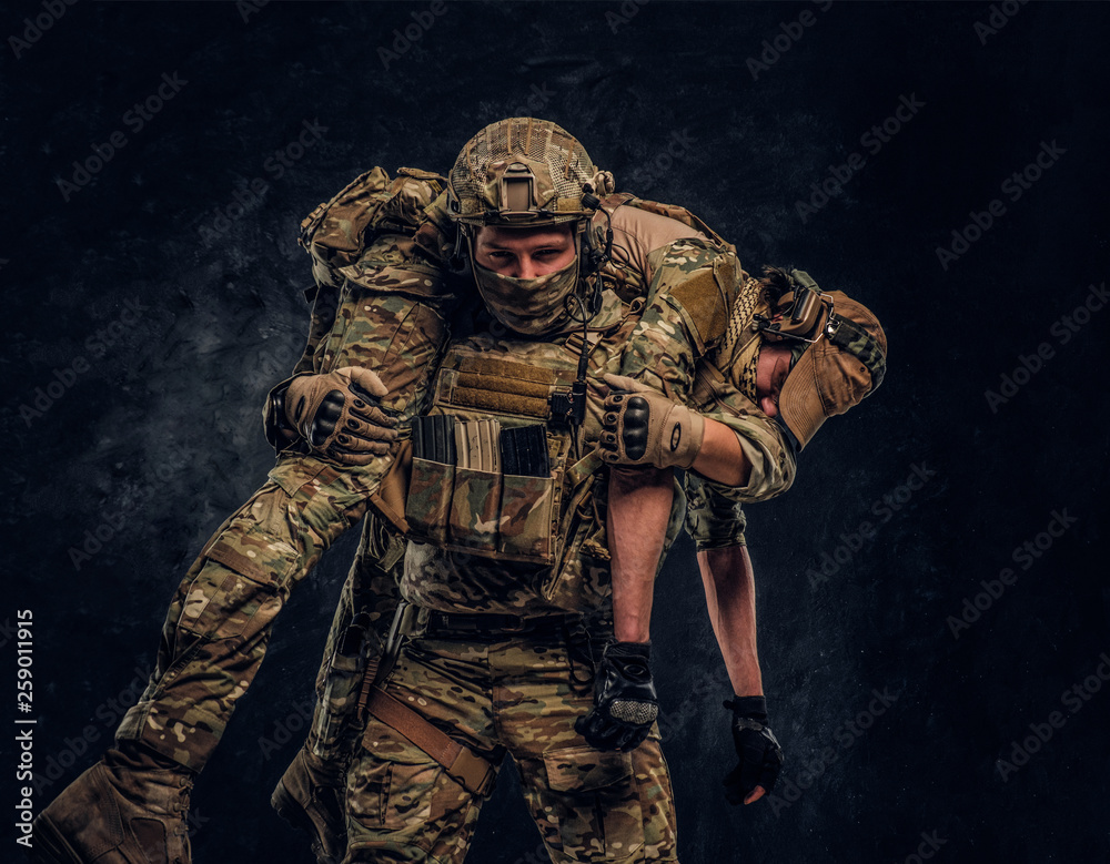Combat conflict, special mission, retreat. Soldier special forces rescue his wounded teammate carrying him on his shoulders from the battlefield. Studio photo against a dark wall