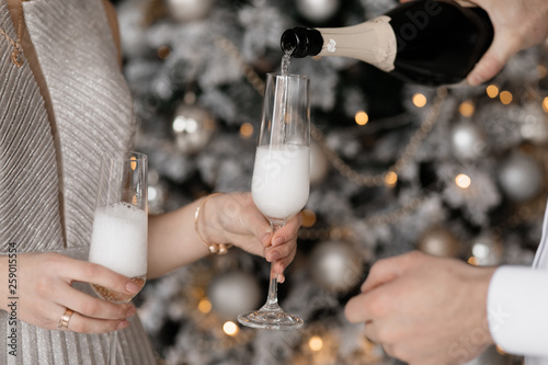 Charming young couple in fancy clothes poses with champagne glasses before a Christmas tree