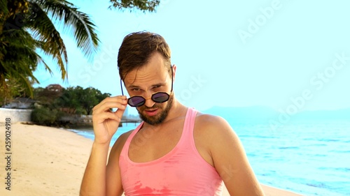 Close-up, Playful handsome guy in pink t-shirt on a tropical beach. he looks at the camera, rejoices and makes funny faces © kopitinphoto