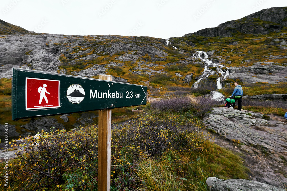 Sign on a hiking trail to the mountain Munkebu on the Lofoten Islands in Norway with a waterfall and a resting young man