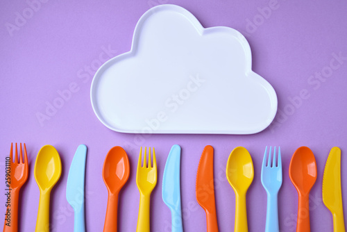 Many colorful plasic forks, spoons and knives with cloud plate on violet background with copy space, top view. photo