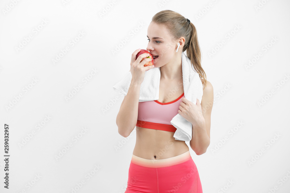 Sports blonde girl in training clothes and with a white towel on his shoulders. He holds in his hand and eats a large red apple in the studio on a white background.