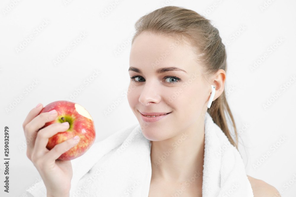 Sports blonde girl in training clothes and with a white towel on his shoulders. He holds in his hand and eats a large red apple in the studio on a white background. Sports blonde girl in training clothes and with a white towel on his shoulders. He holds in his hand and eats a large red apple in the studio on a white background.