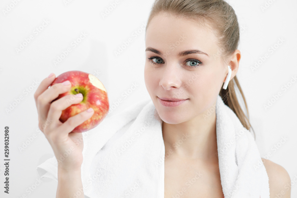 Sports blonde girl in training clothes and with a white towel on his shoulders. He holds in his hand and eats a large red apple in the studio on a white background. Sports blonde girl in training clothes and with a white towel on his shoulders. He holds in his hand and eats a large red apple in the studio on a white background.