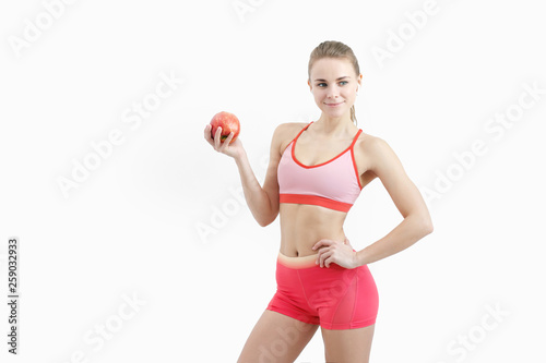 Sports blonde girl in training clothes and with a white towel on his shoulders. He holds in his hand and eats a large red apple in the studio on a white background.