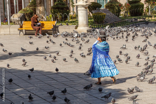 Bolivian woman cholita in blue dress and retro hat walking across La Paz central square full of pigeons, Bolivia