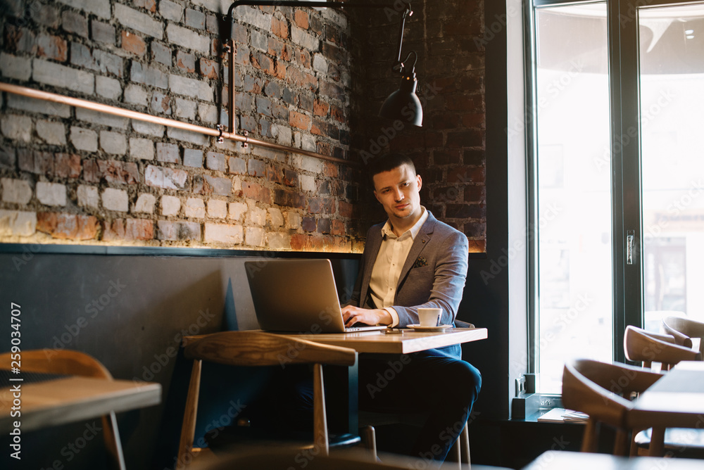 Young handsome man sitting in office with cup of coffee and working on project connected with modern cyber technologies. Businessman with notebook trying to keep deadline in digital marketing sphere. Young handsome man sitting in office with cup of coffee and working on project connected with modern cyber technologies. Businessman with notebook trying to keep deadline in digital marketing sphere.