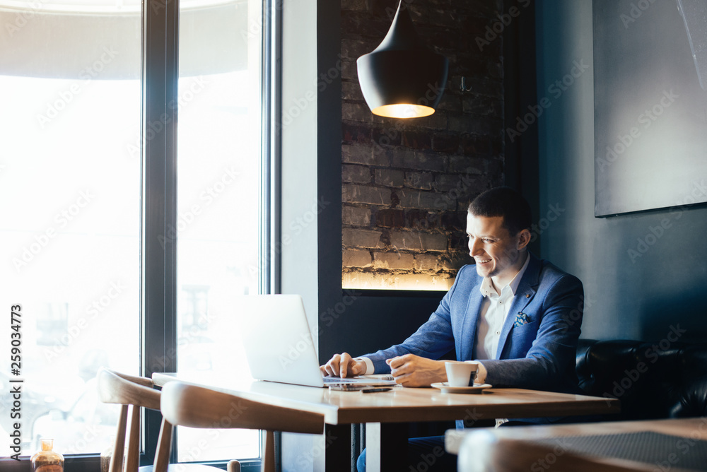 Young handsome man sitting in office with cup of coffee and working on project connected with modern cyber technologies. Businessman with notebook trying to keep deadline in digital marketing sphere. Young handsome man sitting in office with cup of coffee and working on project connected with modern cyber technologies. Businessman with notebook trying to keep deadline in digital marketing sphere.