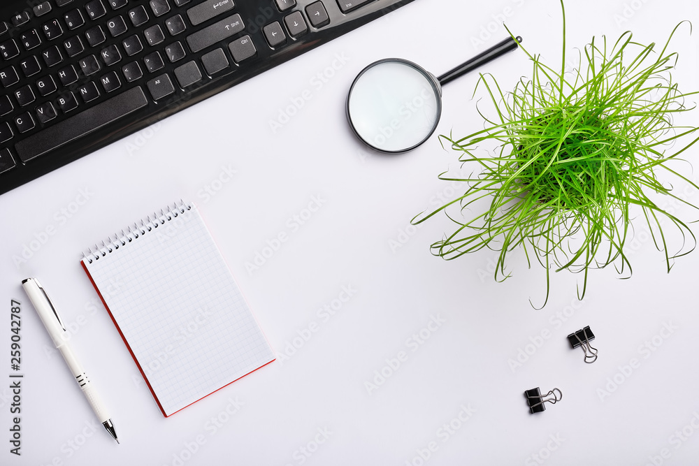 Plakat White surface of the table with keyboard, magnifying glass, notebook, clips, office plant and pen.