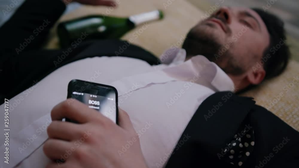 Drunk guy in business clothes laying on bed with smartphone in hand. Boss calling'. Young man still sleeping after party. Monday. Alcohol problems.