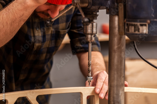 Close up of a man carpenter in a hat and a shirt is carving a wooden board on a large drilling machine in a workshop side view, in the background a lot of tools