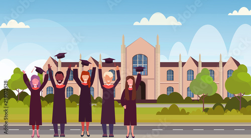 group of mix race students in gown throwing graduation caps in the air standing at campus yard in front of university building exterior education concept flat horizontal full length