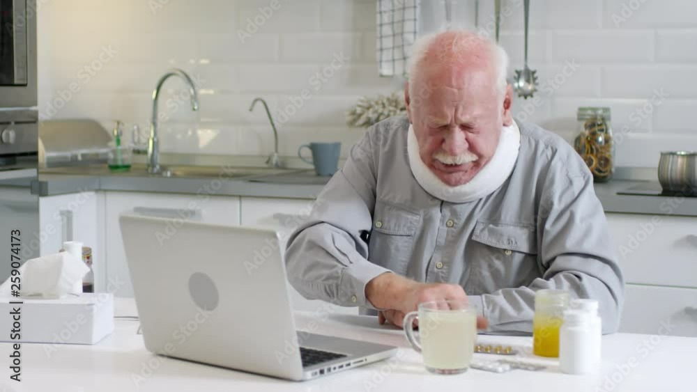 Medium shot of aged Caucasian man in neck bandage sitting at kitchen table, looking at laptop screen and discussing medicines with online physician Medium shot of aged Caucasian man in neck bandage sitting at kitchen table, looking at laptop screen and discussing medicines with online physician