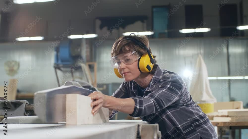 Medium shot of female joiner wearing safety headphones and goggles cutting piece of wood with electric jointer