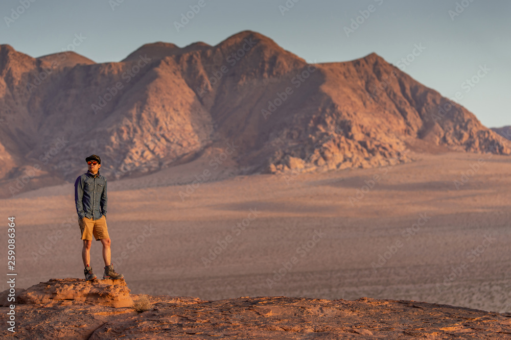 Young Asian man traveler standing on the rock in Wadi Rum desert looking at sunset, famous place in Jordan. Middle east travel concept