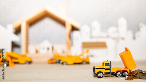 Miniature yellow truck model spilling gold coins on wooden table with blurred house frame and city in the background. Architecture and construction industry concept