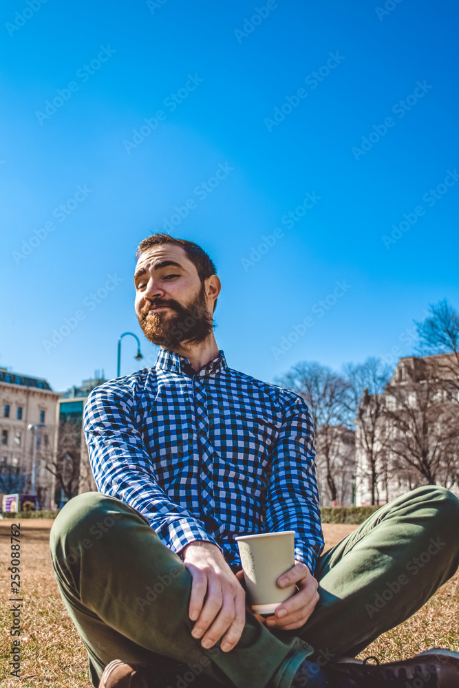 Fototapeta premium The young guy with beard and in shirt sits on grass having crossed legs in the spring park and drinks coffee from paper glass. Mood.