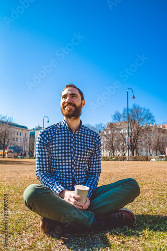 The young guy with beard and in shirt sits on grass having crossed legs in the spring park and drinks coffee from paper glass. Mood.