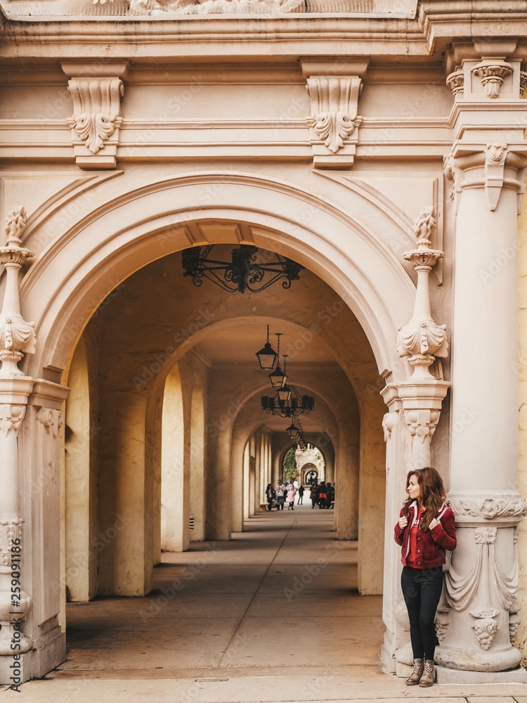 Brunette girl in a red jacket and with a backpack stands near the arch with beautiful stucco and architecture in the Park in San Diego
