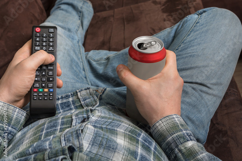 Guy is lying on sofa with TV remote control and drinking beer