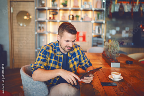 Smiling handsome Caucasian man in plaid shirt sitting in cafe and using tablet. On desk smart phone and coffee.
