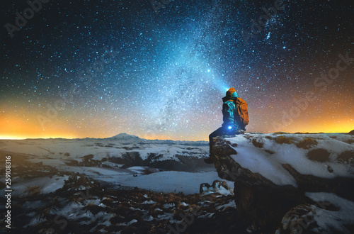 Night winter landscape a man with a backpack and a lantern on his head sits on a rock in the mountains in winter against the background of a mountain and a winter starry sky and the Milky Way