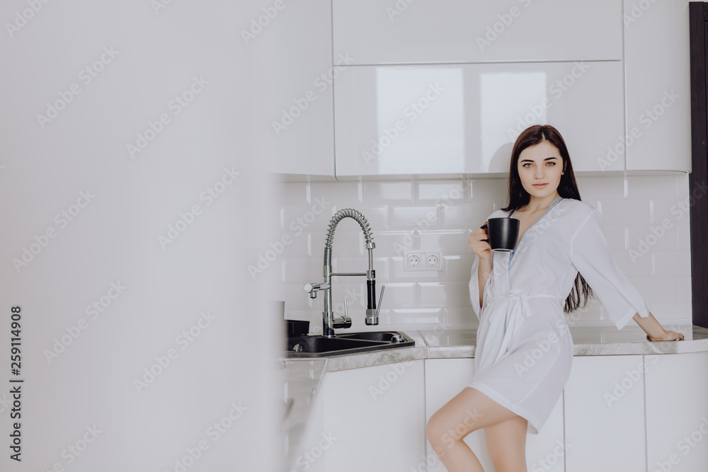 Elegant smiling woman in white robe standing in front of a kitchen drinking an early morning cup of coffee Elegant smiling woman in white robe standing in front of a kitchen drinking an early morning cup of coffee
