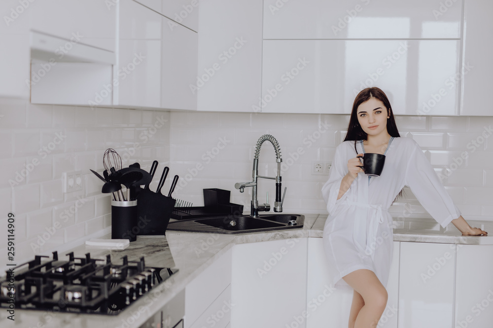Portrait of young woman in white robe after bath holds a cup with coffee or tea against kitchen background. Close up face toothy smile. Portrait of young woman in white robe after bath holds a cup with coffee or tea against kitchen background. Close up face toothy smile.