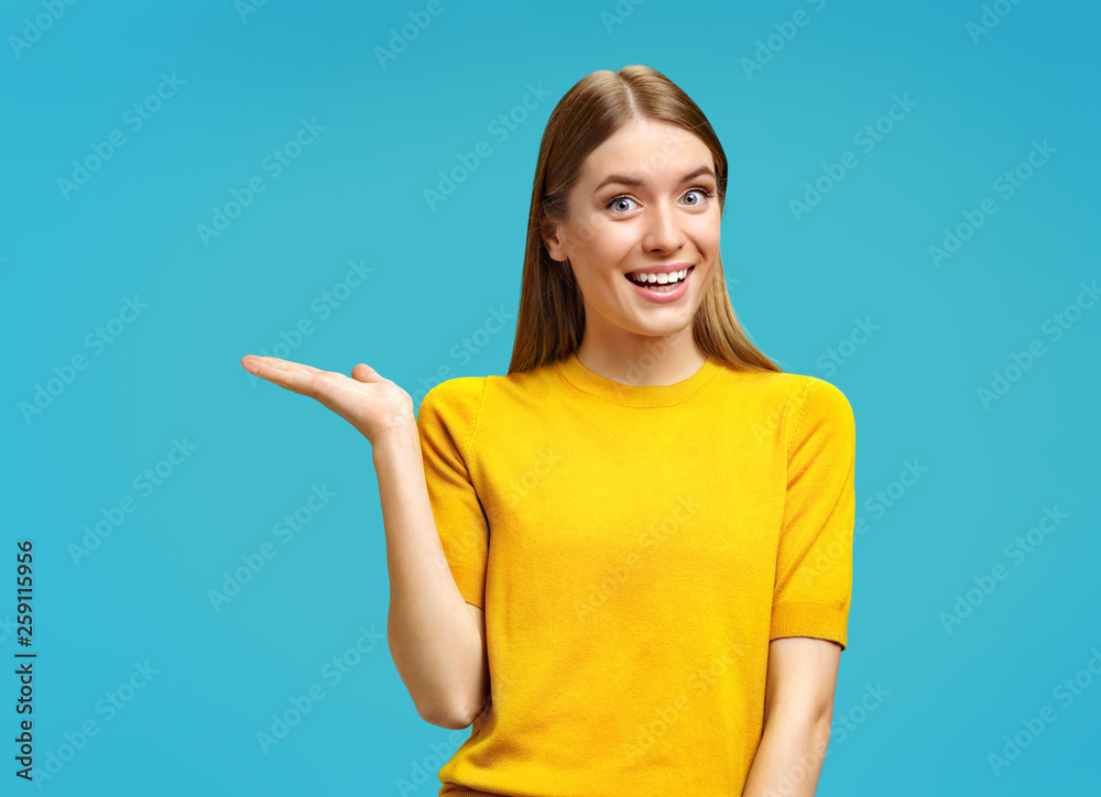 Smiling young girl points aside her open palm, shows copy space for your advert. Photo of attractive girl in yellow sweater on blue background. Smiling young girl points aside her open palm, shows copy space for your advert. Photo of attractive girl in yellow sweater on blue background.