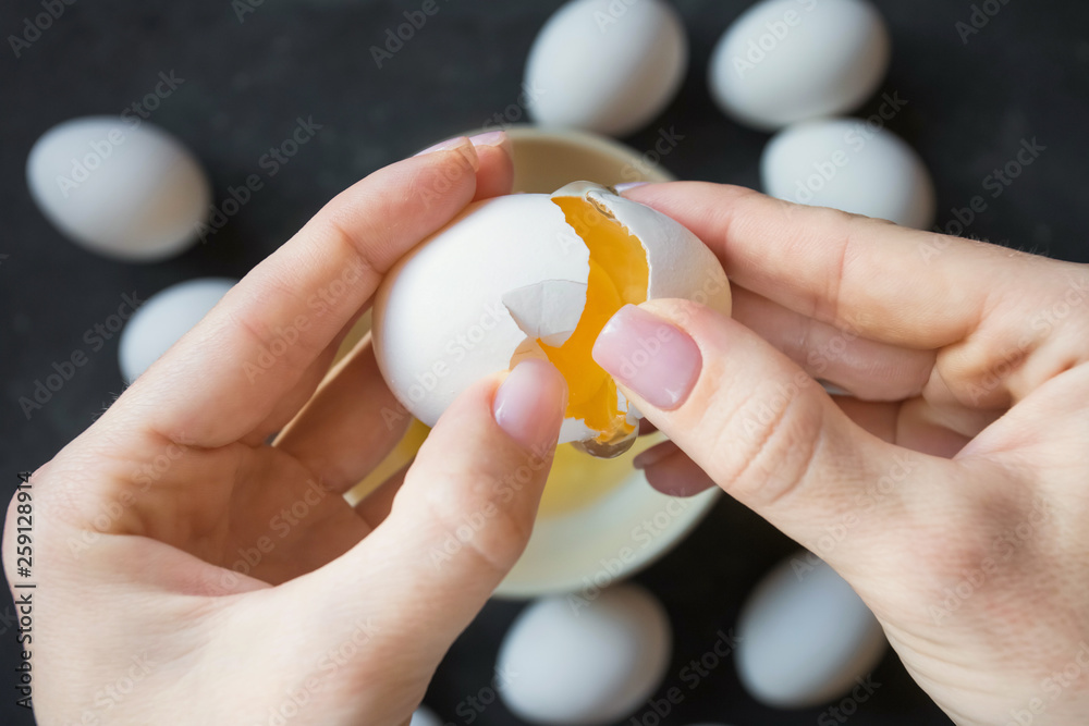 female hands open the shells of a fresh white chicken egg on a dark background with other eggs and yolk and white are visible female hands open the shells of a fresh white chicken egg on a dark background with other eggs and yolk and white are visible