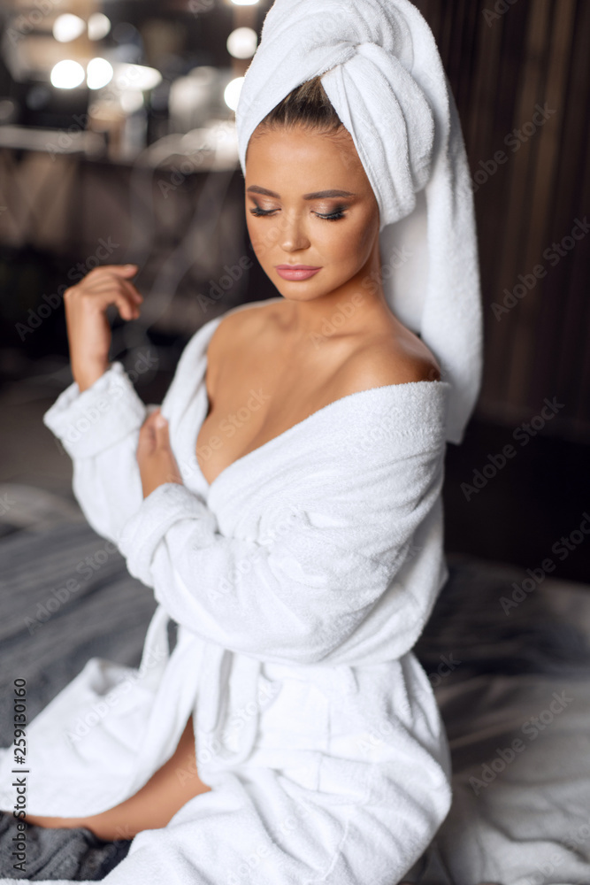 Fototapeta premium Sexy brunette in white coat and towel relaxes on the bed after a shower. Spa