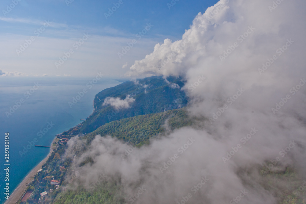 Obraz premium Flying over the bright beautiful green shore and over the blue sea next to the white fluffy clouds. Aerial photo from in the Caucasus in Abkhazia