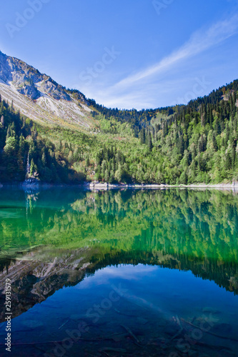 Symmetrical reflections. Turquoise water of a mountain lake surrounded by green wooded hills under a blue sky. Lake Ritsa Tourism in the Caucasus in Abkhazia.