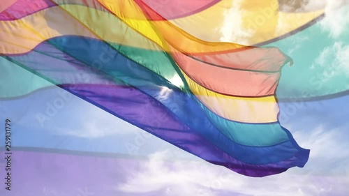 Gay pride rainbow flag flying in slow motion overlay against puffy white clouds in bright blue sky