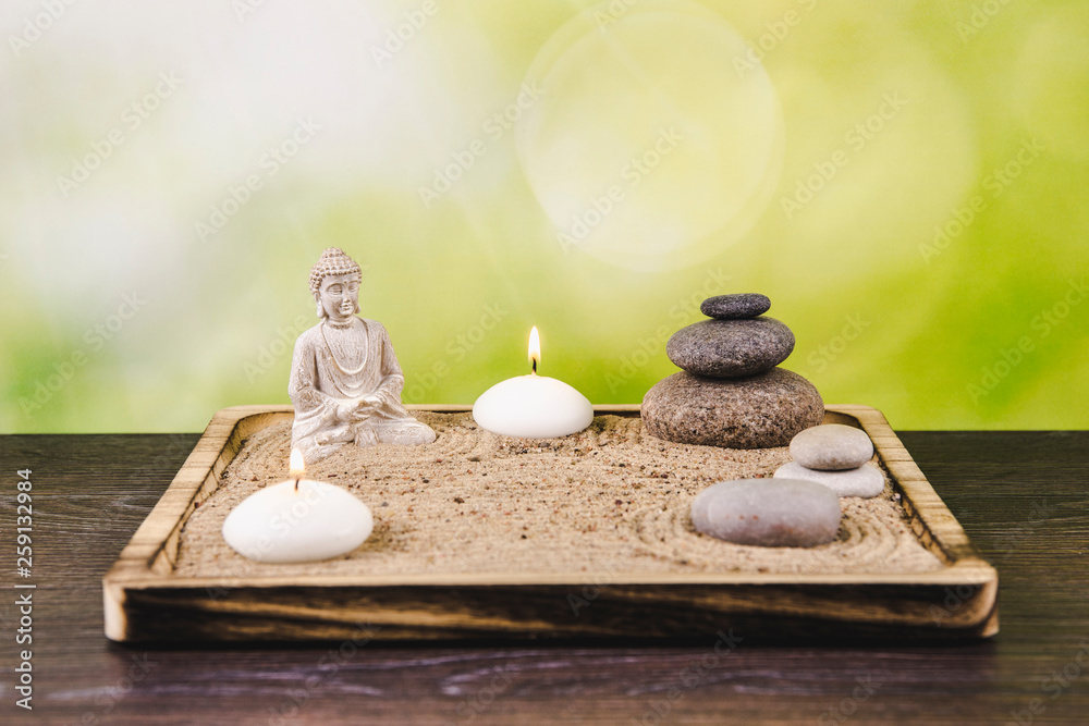 Miniature desk zen sandbox with Buddha figure sit in Lotus position, stacked zen sea stones, spa candles burning against green bokeh studio background, copy space. - obrazy, fototapety, plakaty 