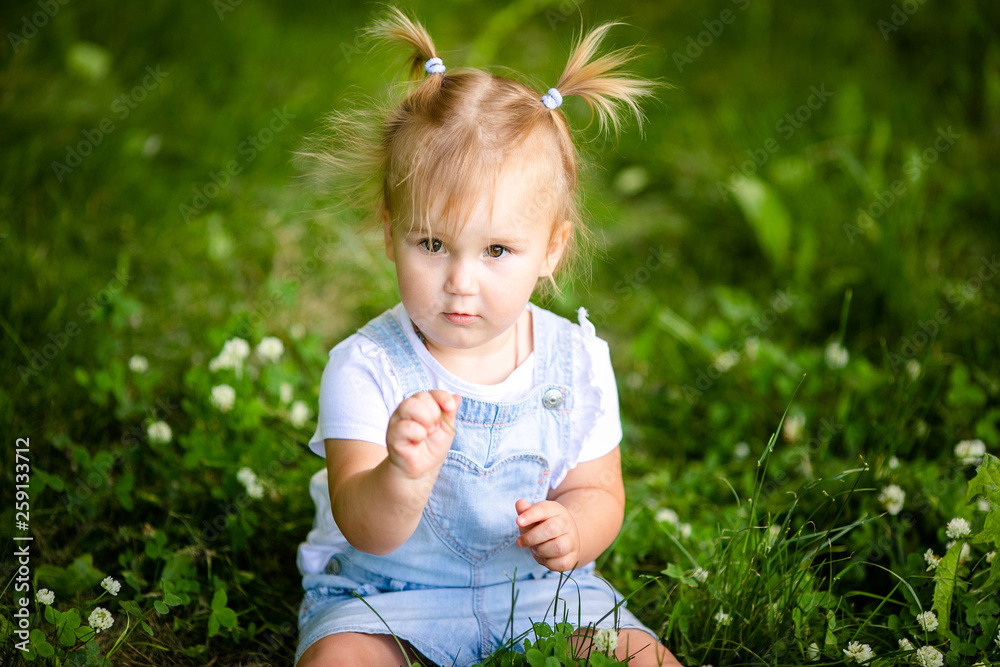 Happy funny blonde baby girl with two little braids in white t-shirt and jeans bodykit sitting on green grass background Happy funny blonde baby girl with two little braids in white t-shirt and jeans bodykit sitting on green grass background
