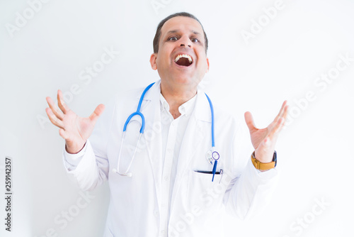Middle age doctor man wearing stethoscope and medical coat over white background celebrating crazy and amazed for success with arms raised and open eyes screaming excited. Winner concept