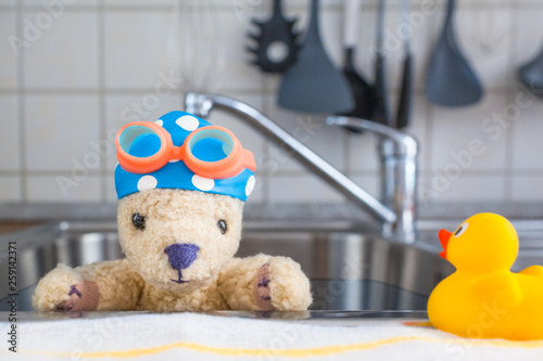 Home Swimming Lesson / Kitchen stainless steel sink as swimming pool, cute toy teddy bear inside wear blue white dotted bathing cap and swim goggles, rubber duck as teacher outside (copy space)