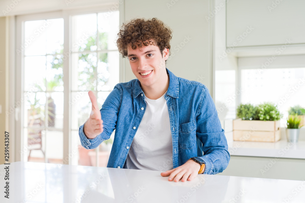 Young handsome man wearing casual denim jacket at home smiling friendly offering handshake as greeting and welcoming. Successful business.