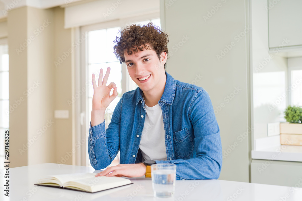Young man reading a book at home doing ok sign with fingers, excellent symbol Young man reading a book at home doing ok sign with fingers, excellent symbol