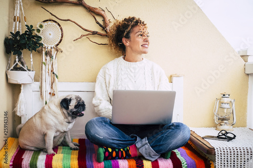 Modern couple with pretty curly woman and funny dog pug sitting on the beanch at home working with a laptop - coloured blanket and girl with technology leisure activity - millennial and love animals