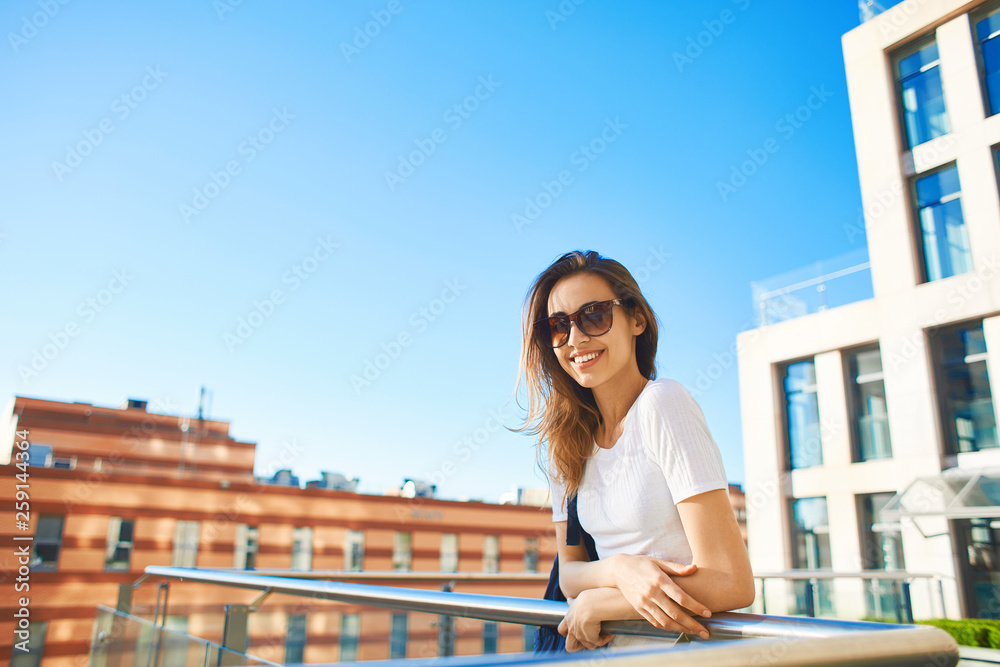 portrait of a young smiling attractive woman in white t-shirt with small city backpack at sunny day on city building and blue sky background. woman poses in cityscape.