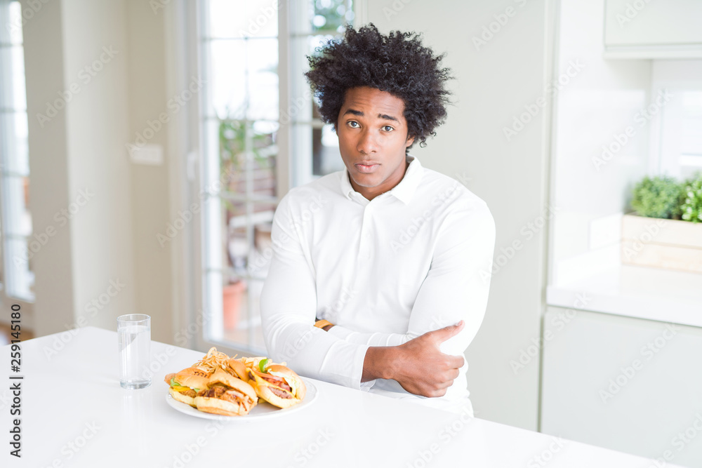African American hungry man eating hamburger for lunch skeptic and nervous, disapproving expression on face with crossed arms. Negative person. African American hungry man eating hamburger for lunch skeptic and nervous, disapproving expression on face with crossed arms. Negative person.