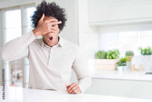 African American man at home peeking in shock covering face and eyes with hand, looking through fingers with embarrassed expression.