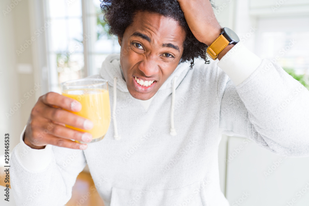 African American man holding and drinking glass of orange juice stressed with hand on head, shocked with shame and surprise face, angry and frustrated. Fear and upset for mistake. African American man holding and drinking glass of orange juice stressed with hand on head, shocked with shame and surprise face, angry and frustrated. Fear and upset for mistake.