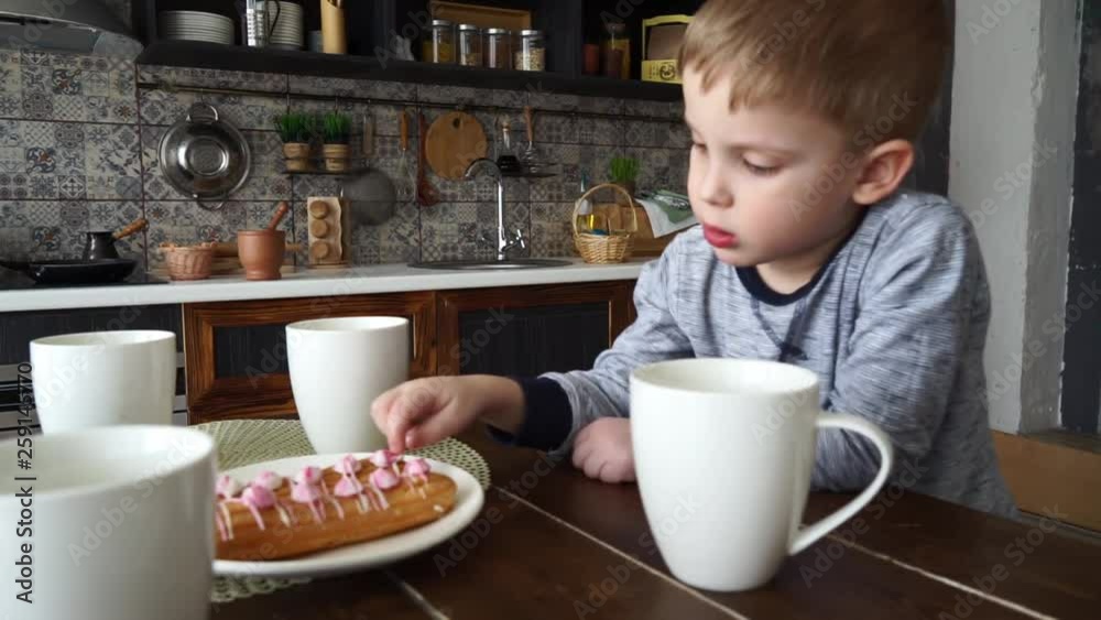 A pensive little boy sits at a table with a circley tea and cakes. Licking his finger. Portrait. A pensive little boy sits at a table with a circley tea and cakes. Licking his finger. Portrait.