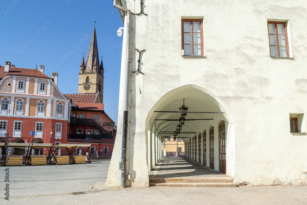 Old, medieval, white building with a corridor with several archways and a church tower in the background, on 'Piata Mica' Square - Sibiu (Hermannstadt), Transylvania, Romania