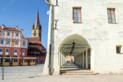 Old, medieval, white building with a corridor with several archways and a church tower in the background, on 'Piata Mica' Square - Sibiu (Hermannstadt), Transylvania, Romania