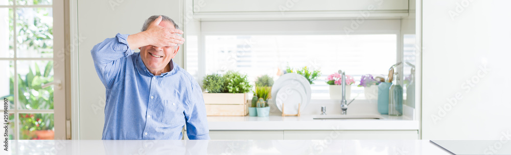 Wide angle perspective of handsome senior man at home smiling and laughing with hand on face covering eyes for surprise. Blind concept. Wide angle perspective of handsome senior man at home smiling and laughing with hand on face covering eyes for surprise. Blind concept.